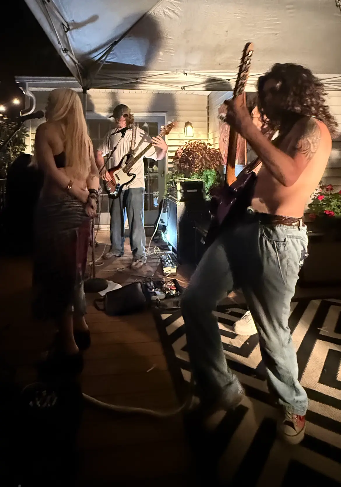The band playing on a back porch at night under a canopy, the guitarist leaning into a solo in the foreground with the bassist and singer behind him